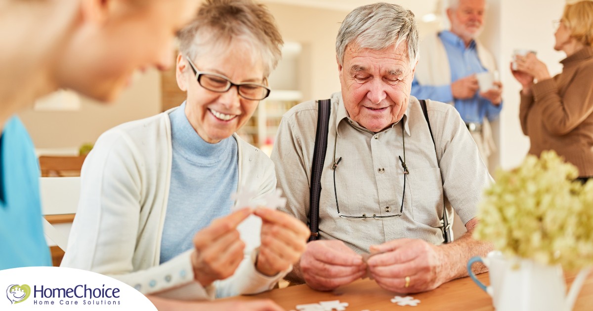 An older couple works on a puzzle with a caregiver, representing the kind of activity that helps those with dementia and also representing Alzheimer’s and Brain Awareness Month.