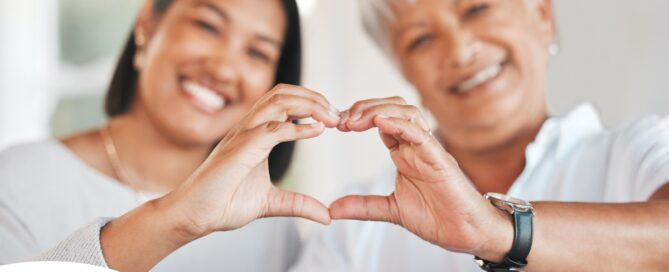 A daughter and an older mother create a heart with their hands, representing the feeling that is highlighted during National Family Caregivers Month