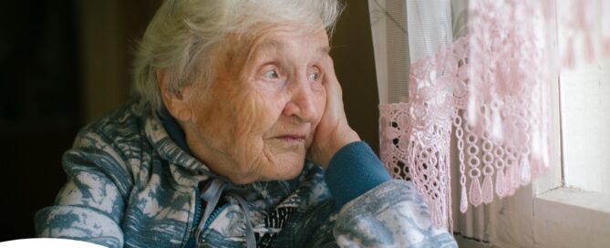 An older woman peacefully looks out of the window while the sun is still up, representing what can happen with sundowning.